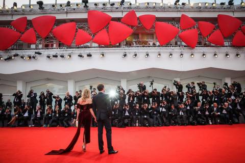 A couple in formal attire walks hand-in-hand on a red carpet, surrounded by a throng of photographers, with a backdrop of red abstract sculptures and tiered lighting.