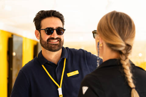 A smiling man in sunglasses and a blue polo shirt talks to a woman with a braided blonde ponytail in the foreground, while a blurred yellow and black background is visible behind them.