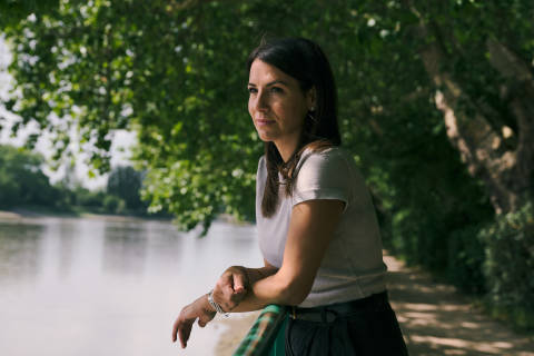A woman leans on a railing in the foreground, looking out at a calm body of water bordered by trees in the background.