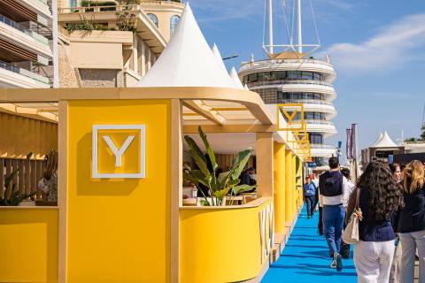 A bright yellow structure with a white "Y" logo stands in the foreground, while people walk along a blue path toward a modern white building under a clear blue sky.