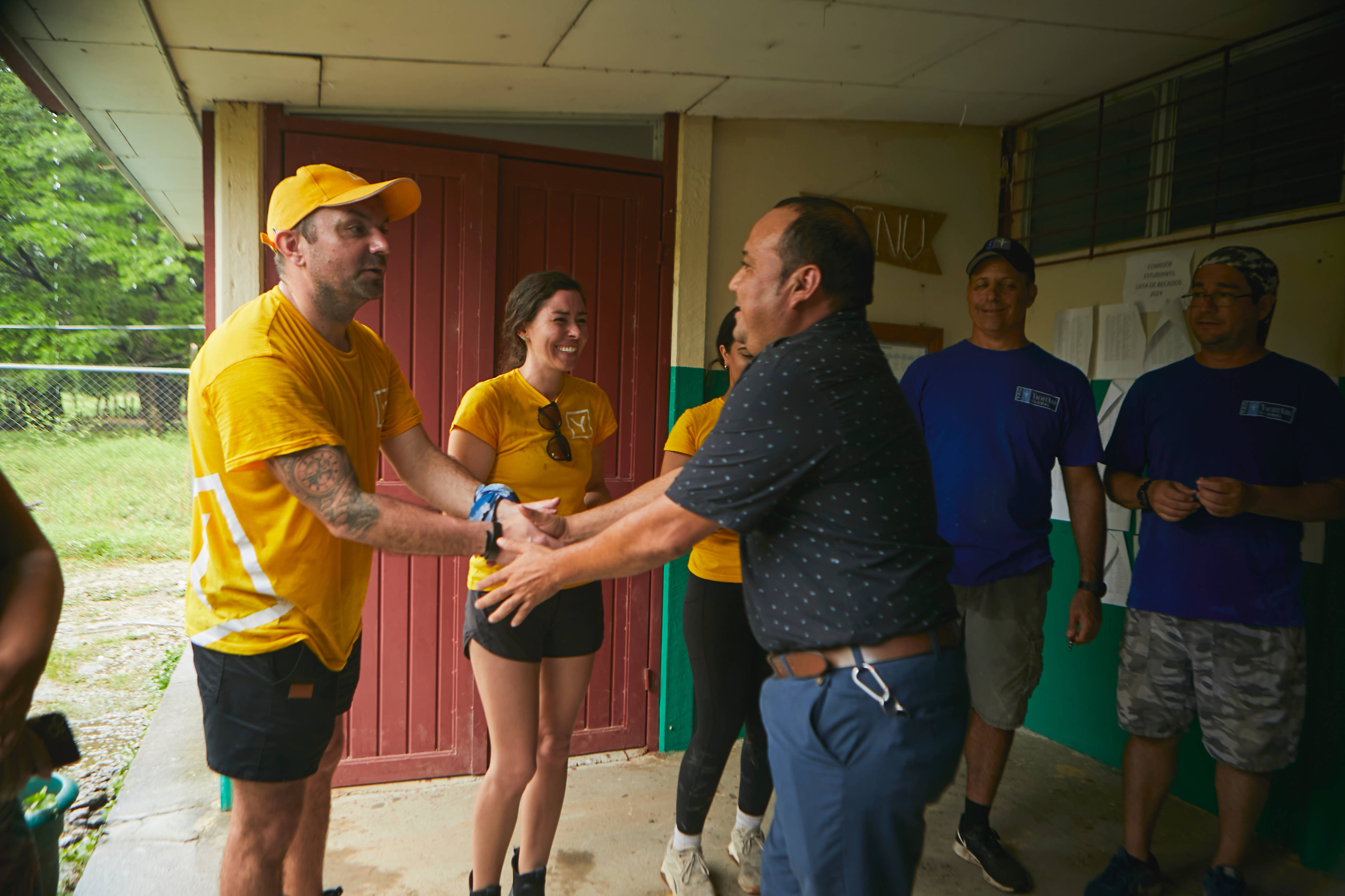 A group of people in yellow and blue shirts are gathered outdoors, some shaking hands in a friendly greeting.