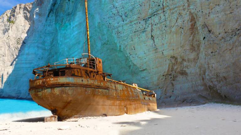 A rusty shipwreck rests on a white sandy beach against a backdrop of towering, turquoise-tinged cliffs.