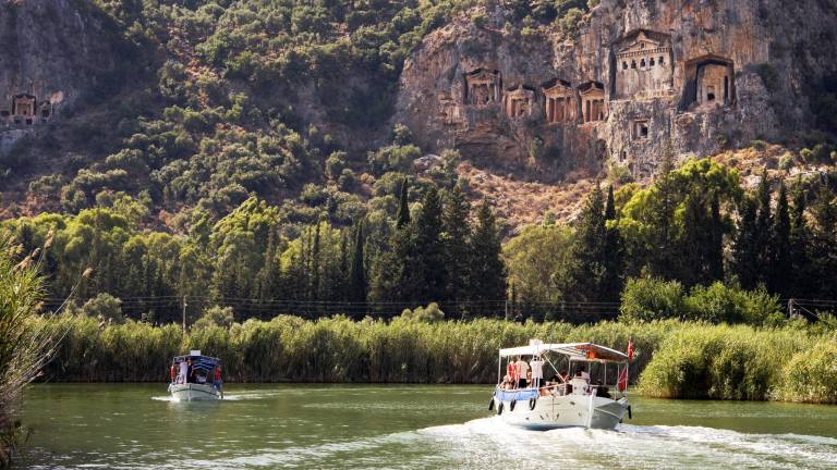 Tourists on a boat cruise past ancient Lycian rock tombs carved into a towering cliff face.