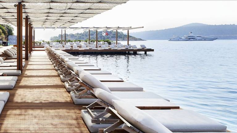 Luxurious lounge chairs line a wooden deck overlooking the tranquil blue water and a distant yacht.