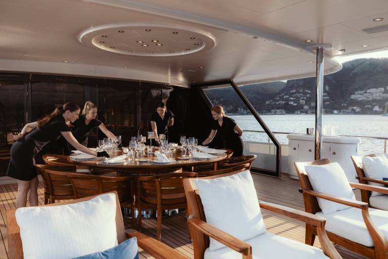 Four servers meticulously set a large dining table on a yacht's deck, overlooking a picturesque coastal town.