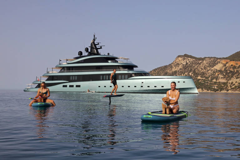 A luxurious yacht serves as a backdrop for people enjoying water sports and their dogs on a sunny day.