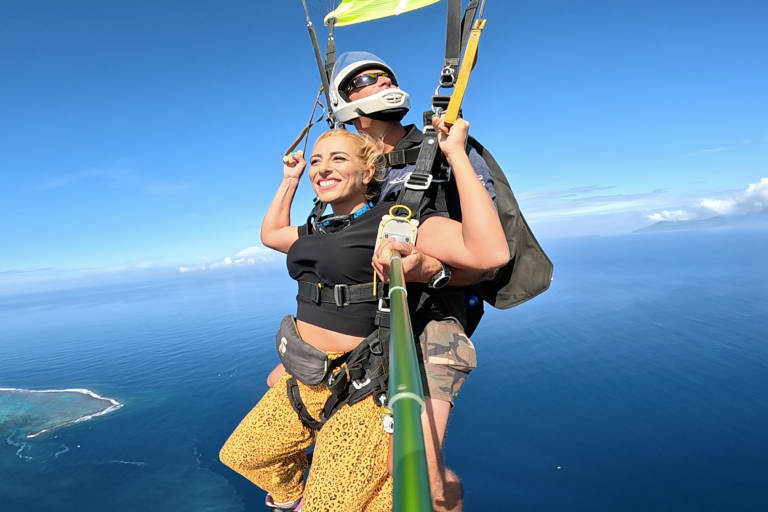 A smiling woman in leopard print pants enjoys a tandem skydive over a tropical ocean with a distant island.