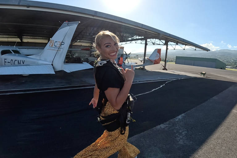 A smiling woman in a leopard print dress and parachute harness walks on an airfield near a small airplane.