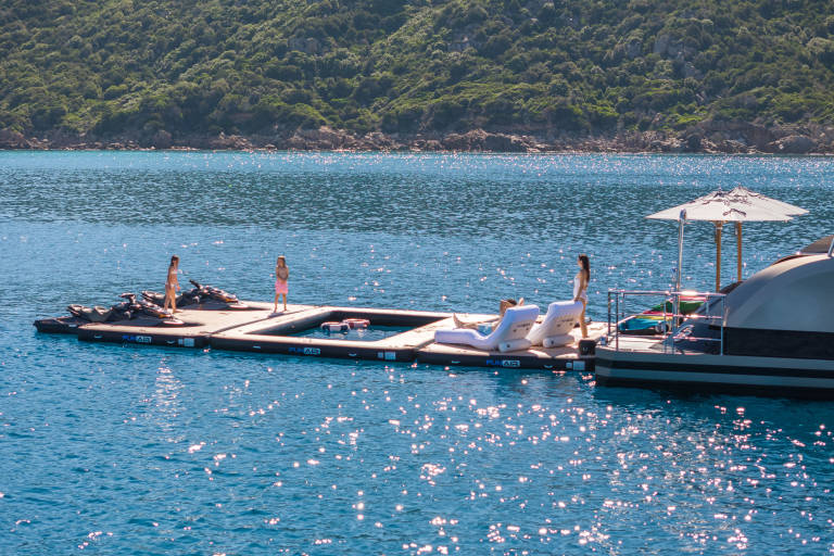 People enjoy a sunny day on a floating dock with jet skis and lounge chairs, anchored near a lush green coastline.