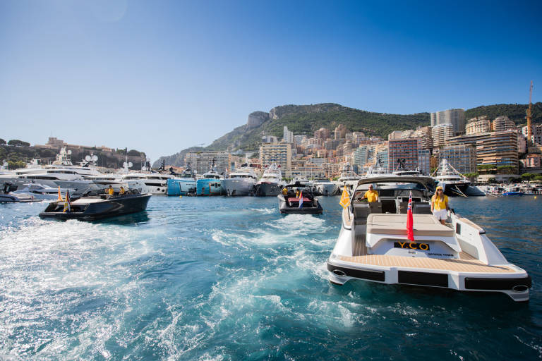 Luxury yachts navigate the sparkling blue waters of a Mediterranean harbor with a picturesque city skyline in the background.