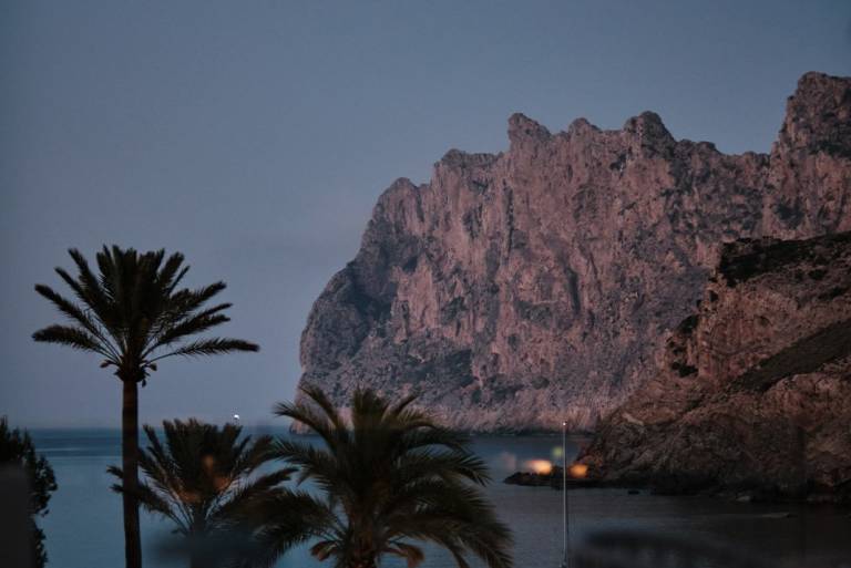 Towering rocky cliffs rise against a clear sky, with palm trees in the foreground framing the dramatic landscape.