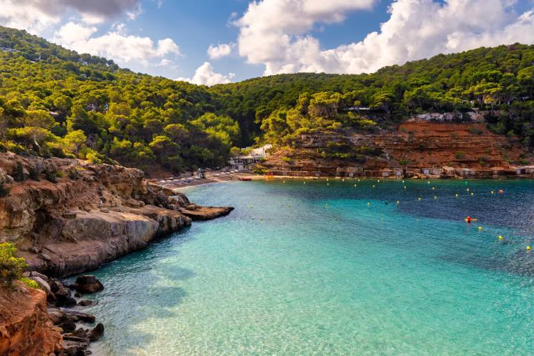 Rocky cliffs and a small beach border the crystal-clear turquoise water, with lush green hills and a bright blue sky forming the background.
