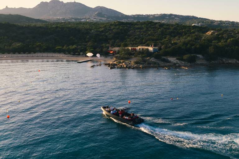 A boat with people on board cruises across the blue water in the foreground, with a tree-covered coastline and hills in the background.