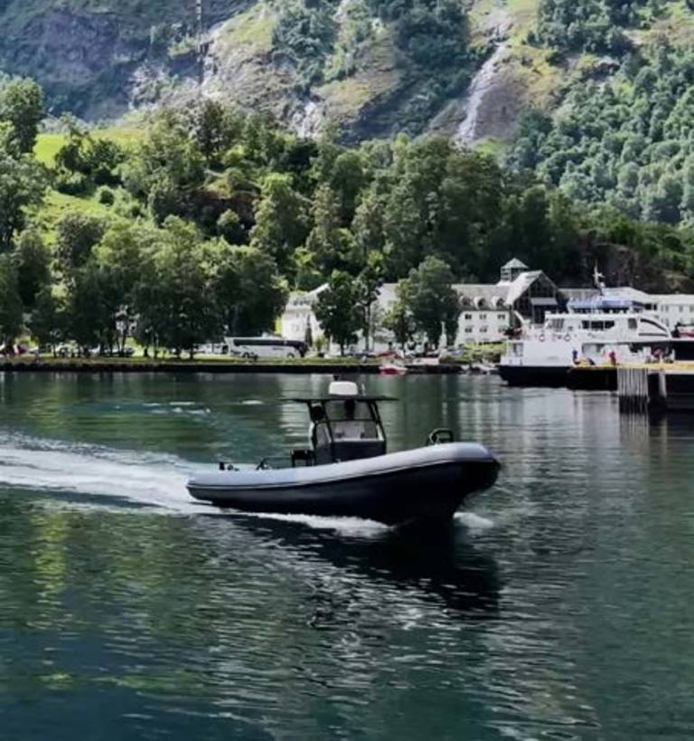 A dark inflatable boat speeds across calm water in the foreground, with a village nestled at the base of a lush, green mountain in the background.