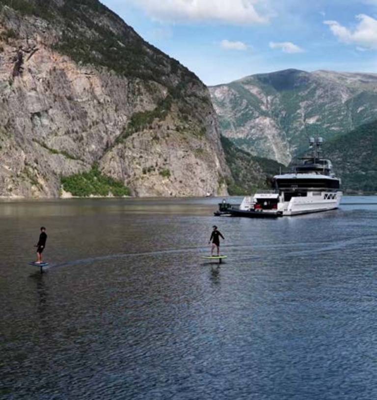 Two people on hydrofoils glide across a calm lake in the foreground, with a large white yacht anchored in the background against a backdrop of steep, tree-covered mountains.