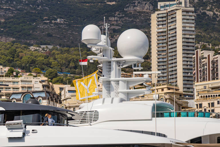 A white luxury yacht with large radar domes stands in the foreground against a backdrop of tall buildings and a steep, forested mountain.