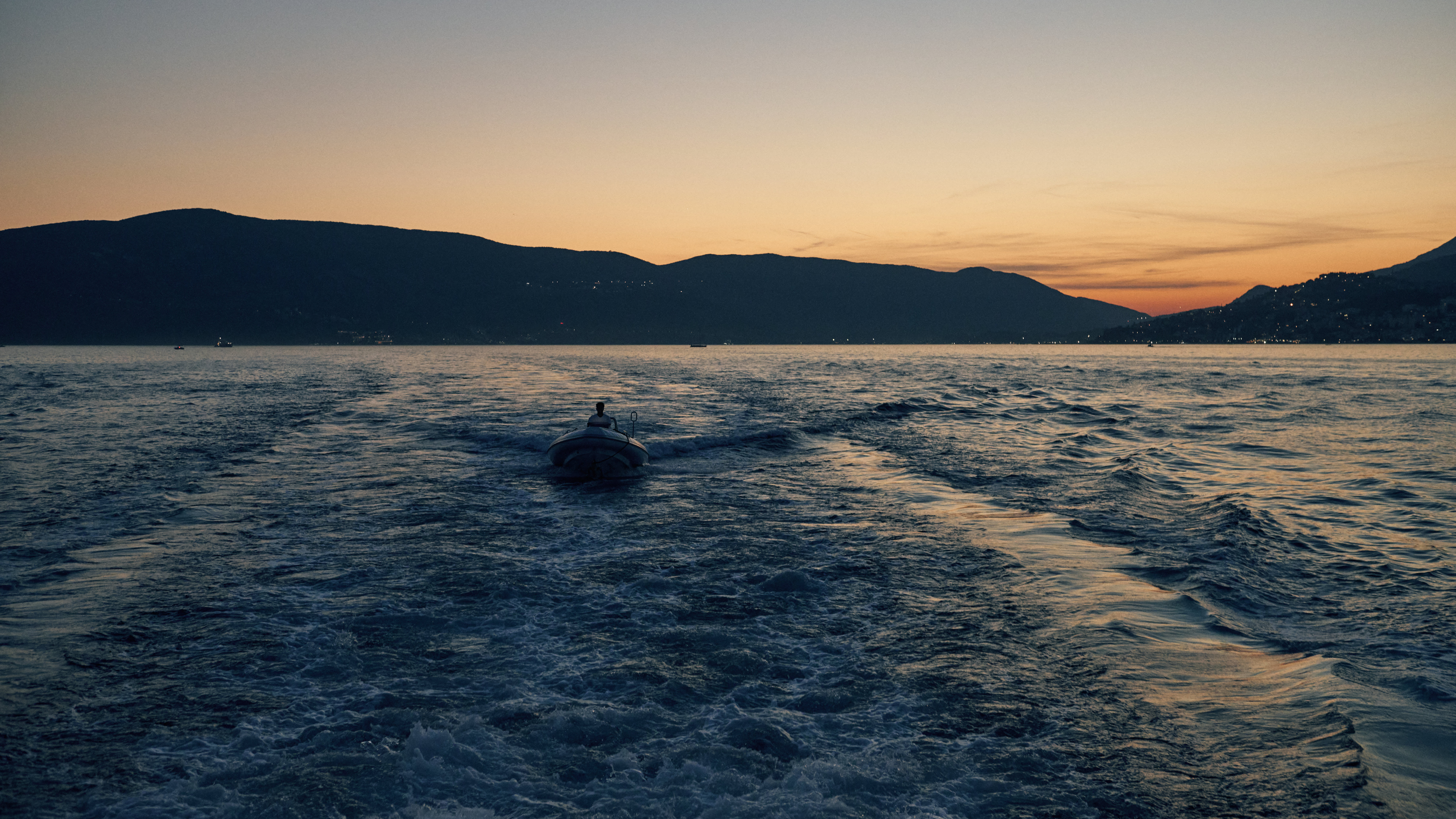 A small boat glides across the calm waters of a lake, surrounded by majestic mountains silhouetted against a vibrant sunset sky.