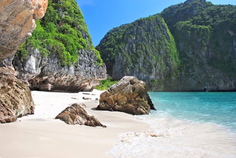 Turquoise waters gently lap the pristine white sand of this secluded tropical cove, framed by lush green cliffs.