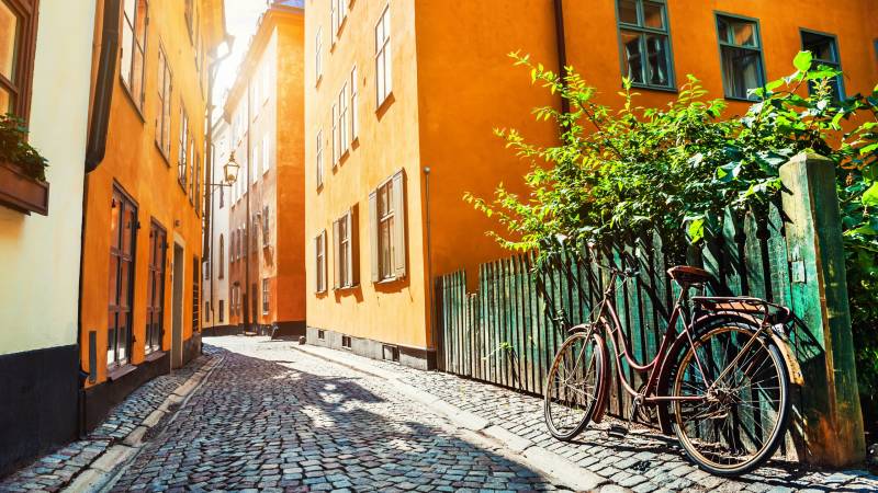 A vintage bicycle leans against a weathered fence on a sun-drenched cobblestone street lined with colorful buildings.