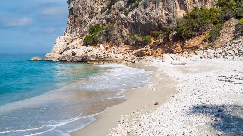 Turquoise waves gently lap a pebble-strewn beach at the base of a rugged, verdant cliff.