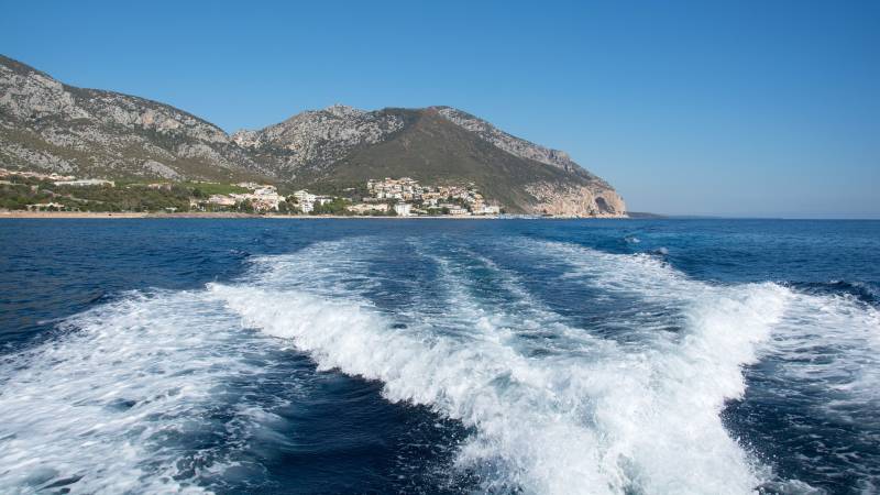 A boat's wake cuts through the deep blue sea as a coastal town nestles against a rugged, verdant mountain.