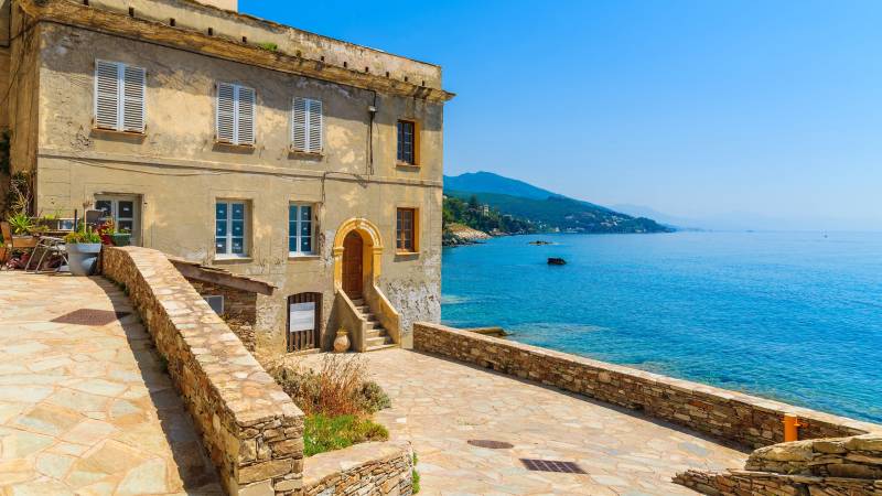 A weathered building overlooks the sparkling blue sea and distant green hills under a clear sky.