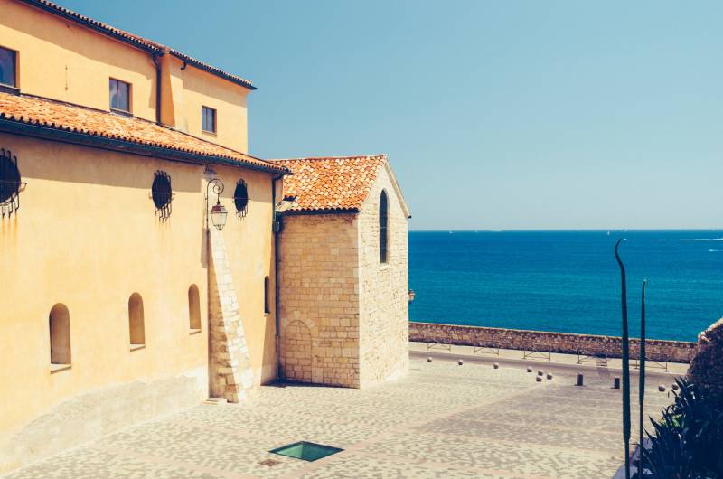 A sun-drenched building with terracotta roofs overlooks the vibrant blue sea under a clear sky.