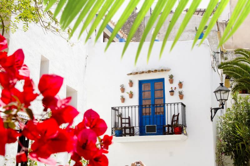 Bright red bougainvillea frames a charming balcony with a vibrant blue door, overlooking a sun-drenched Mediterranean courtyard.