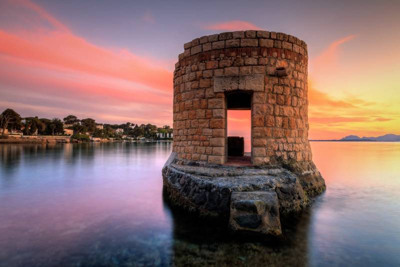 A stone tower stands sentinel over tranquil waters as a vibrant sunset paints the sky with hues of pink and orange.