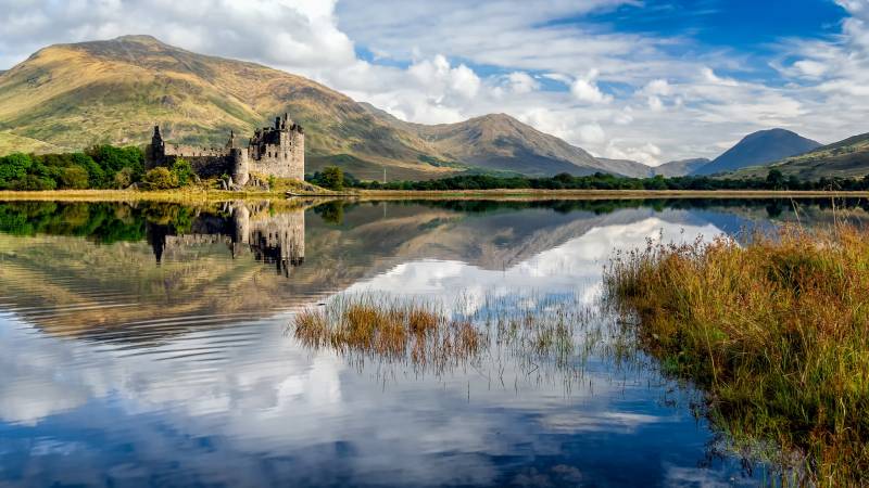Kilchurn Castle stands majestically on a peninsula in Loch Awe, its ruins reflected in the tranquil, rippling water under a dramatic sky.