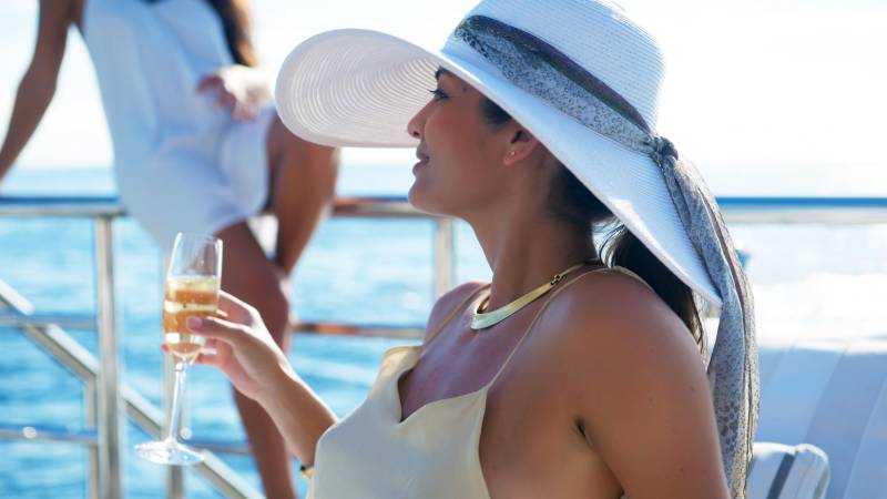 A woman in a wide-brimmed hat sips champagne while enjoying a sunny day on a yacht.