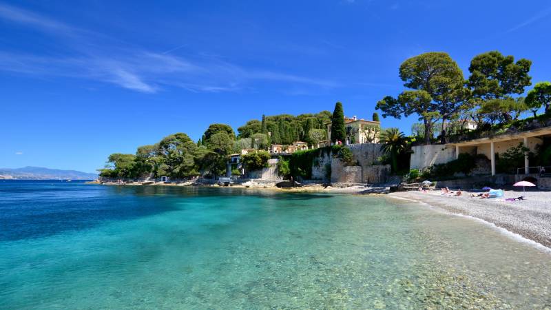 Crystal-clear turquoise waters lap a pebble beach lined with lush greenery and elegant villas under a brilliant blue sky.