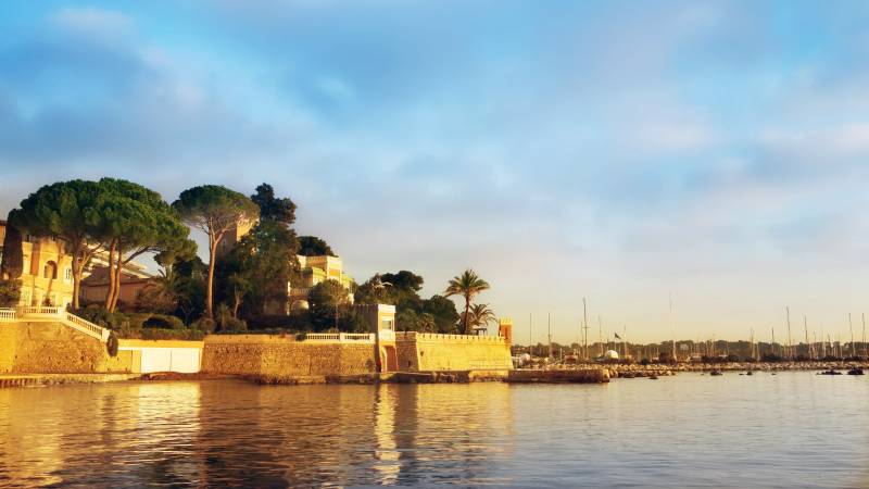 Golden hour light bathes a coastal villa and marina, reflecting on the tranquil Mediterranean waters.