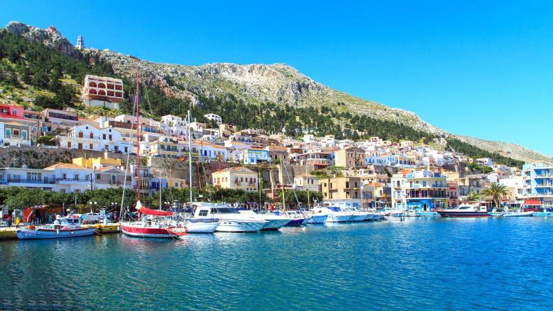Colorful buildings climb a steep hillside overlooking a harbor filled with sailboats and motorboats under a bright blue sky.