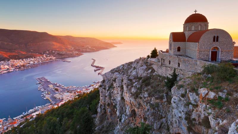 A picturesque Greek Orthodox church overlooks a bustling harbor at sunset.