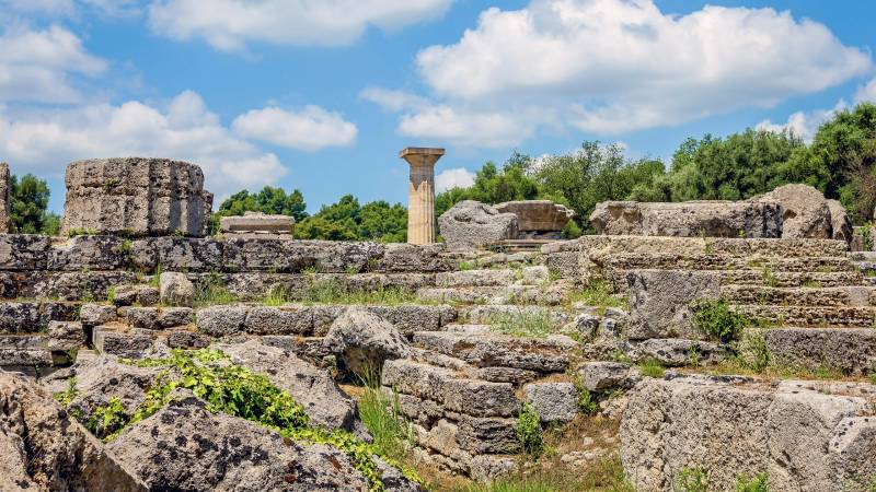 Ancient ruins of stone columns and structures stand amidst lush greenery under a bright blue sky.