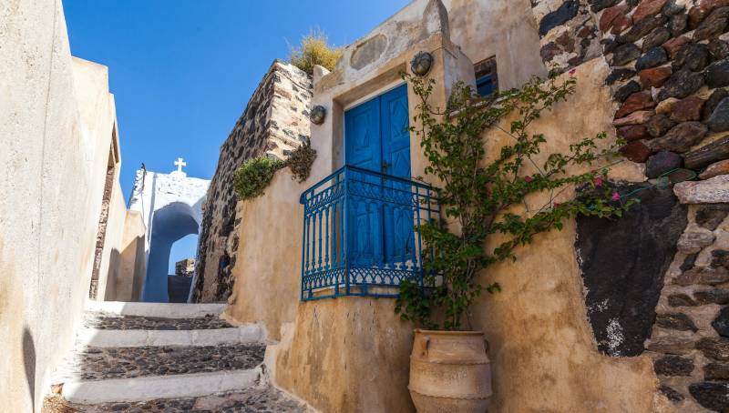 A charming pathway winds past a traditional stone building with a vibrant blue door and balcony in Santorini.