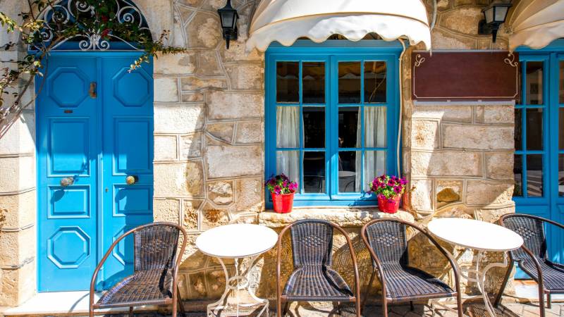 A vibrant blue door and window frame a charming cafe with wicker chairs and tables set against a rustic stone wall.