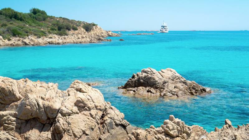 A luxurious yacht cruises on the crystal-clear turquoise waters of a Mediterranean bay fringed by rocky shores.