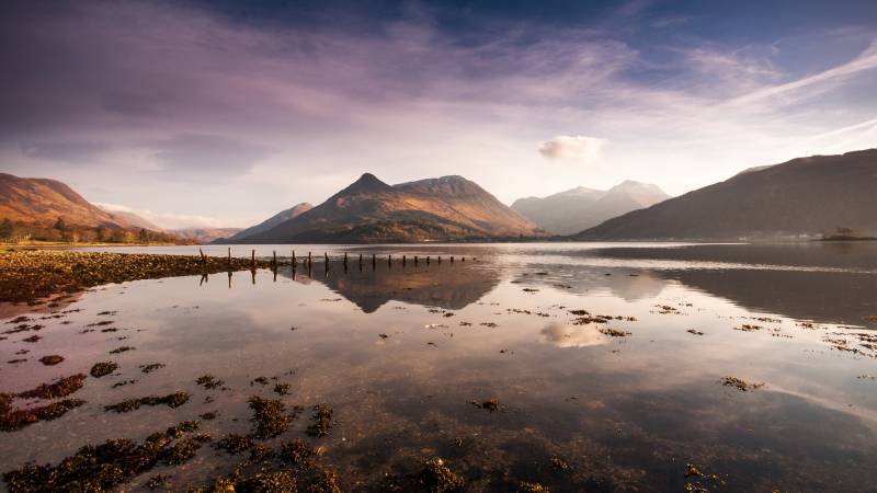Golden mountains reflect in a calm loch under a dramatic purple sky.
