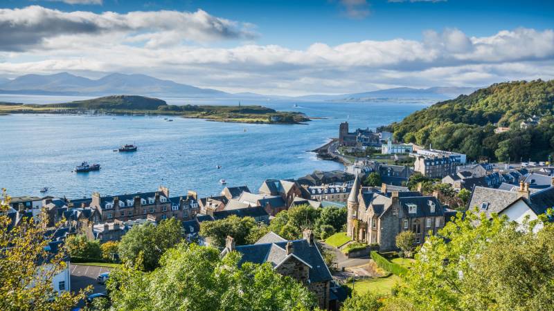 A charming coastal town with historic buildings and a bustling harbor is nestled against a backdrop of rolling green hills.