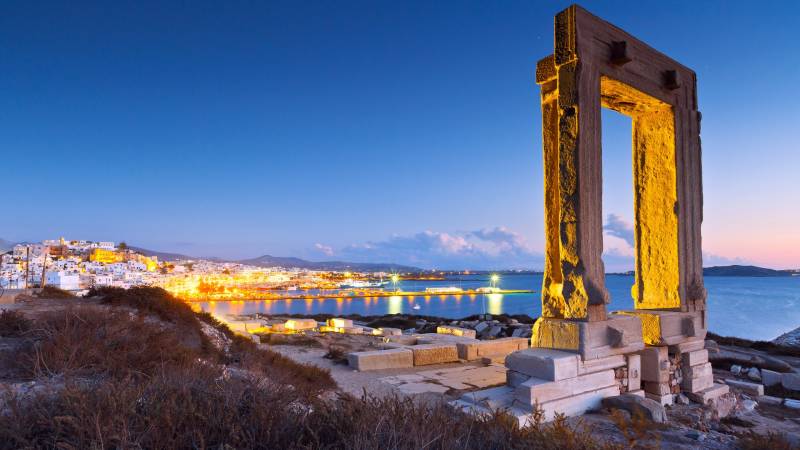Ancient ruins stand illuminated against the twilight sky overlooking a vibrant harbor town.