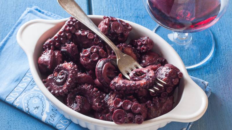 A fork pierces tender, braised octopus tentacles served in a white dish alongside a glass of red wine.