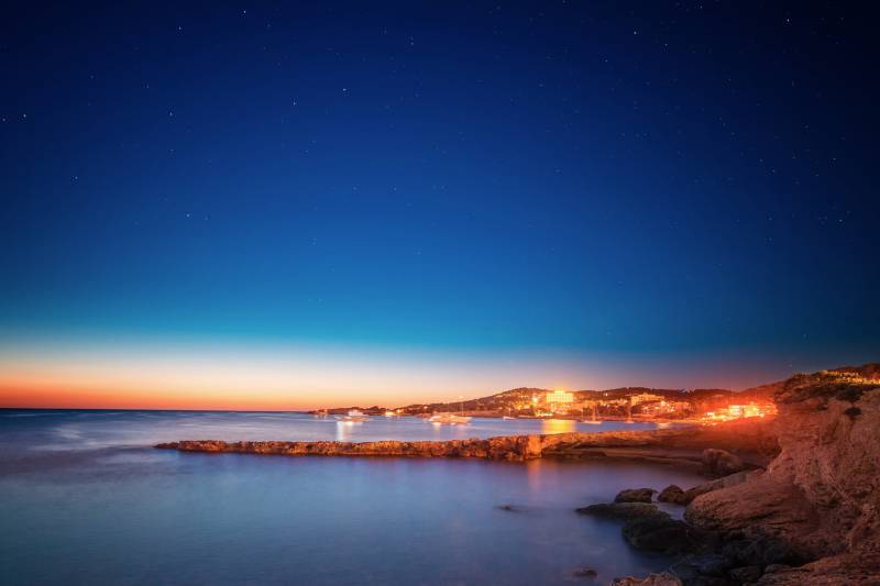 Stars twinkle above a tranquil bay as the lights of a coastal town begin to glow at twilight.