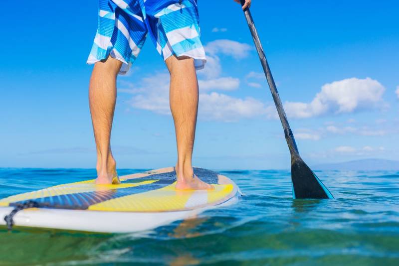 A person in blue and white striped shorts stands on a paddleboard, holding a paddle in the clear blue ocean.