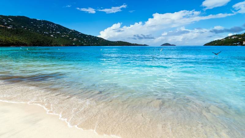 Turquoise waters gently lap onto a pristine white sand beach, with lush green hills dotted with houses rising in the background under a bright blue sky.