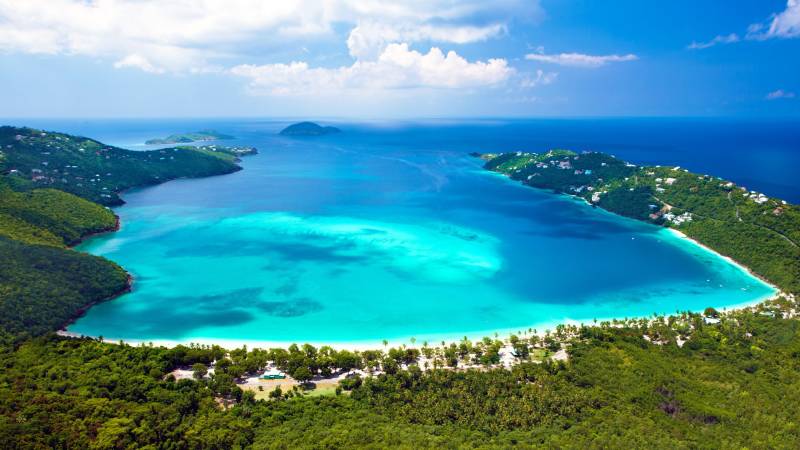 Lush green hills dotted with white homes cascade down to a breathtaking turquoise bay under a bright blue sky.