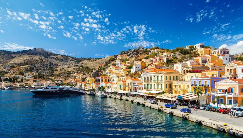 Colorful buildings line the harbor of a Greek island, with a large ferry docked and boats bobbing in the bright blue water.