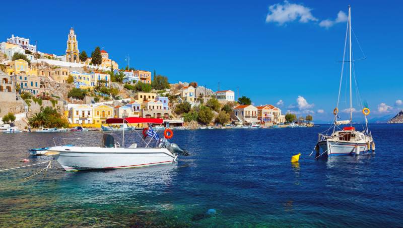 Colorful buildings cascade down a hillside overlooking a tranquil blue bay dotted with boats and a sailboat.