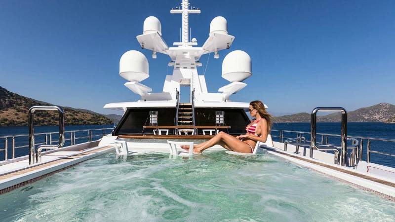 A woman in a striped bikini relaxes in a bubbling hot tub on the deck of a luxurious yacht, surrounded by the azure sea and rugged coastline.